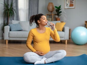 Pregnant woman sitting on a yoga mat drinking water Pregnant woman sitting on a yoga mat drinking water