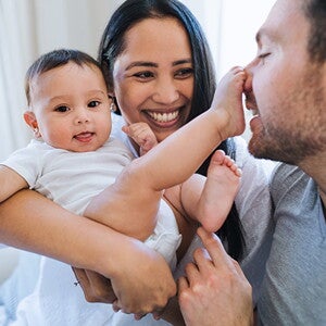 Woman holding her baby while they play with mans face with feet