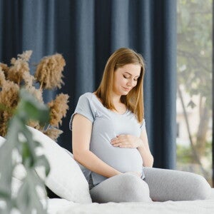 Pregnant woman in grey top and pants sitting on her bed holding her belly