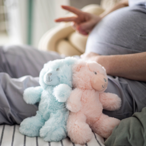 a blue and pink teddy bear and a pregnant woman showing two fingers