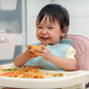 Toddler girl holding corn on a messy highchair
