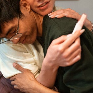 man in green shirt and woman in white shirt hugging with a pregnancy stick in hand