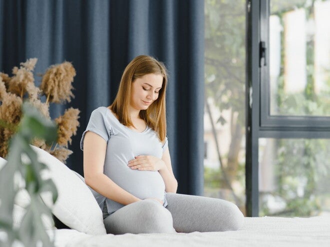 Pregnant woman in grey top and pants sitting on her bed holding her belly Pregnant woman in grey top and pants sitting on her bed holding her belly